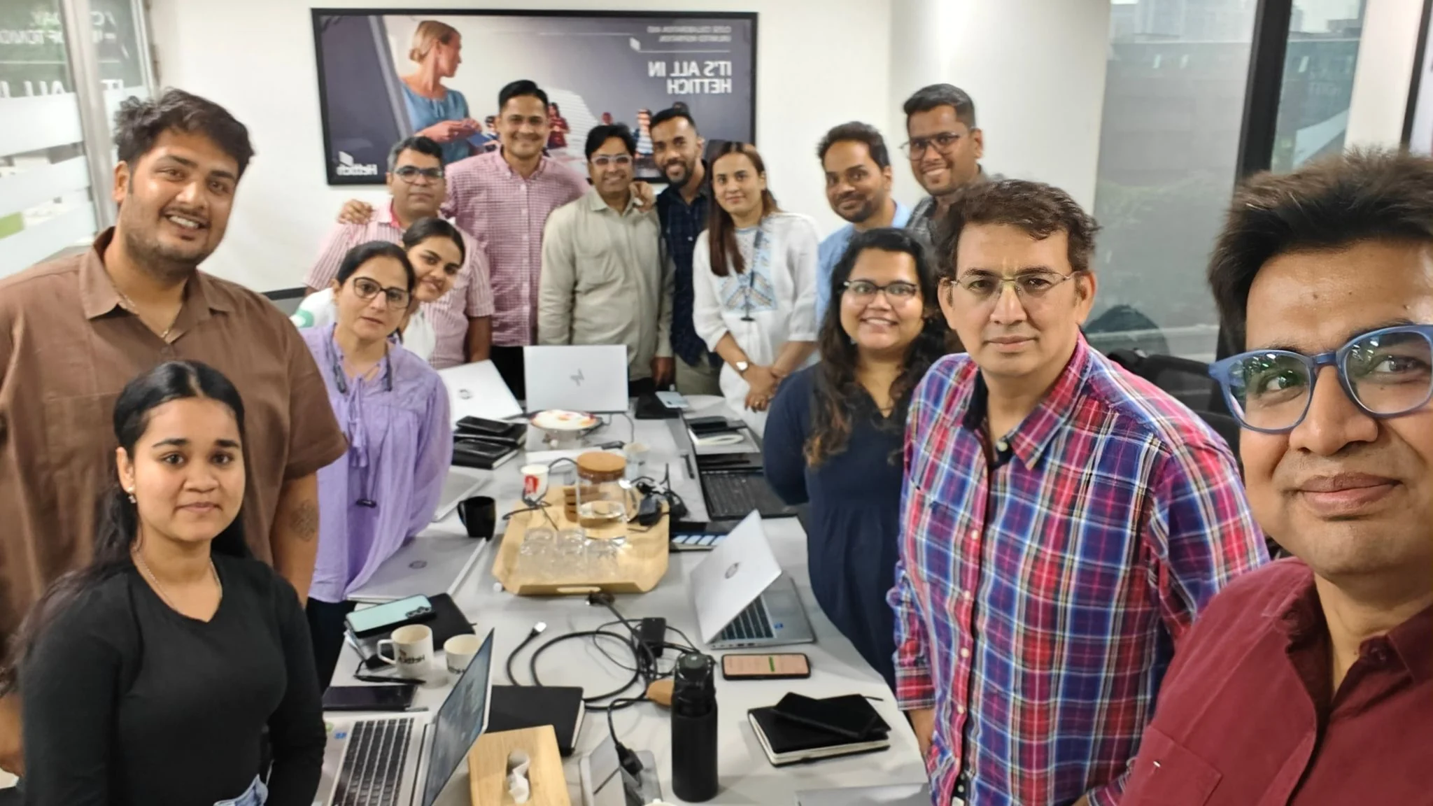 The Brand Vibe AI workshop group at Hettich India: around a dozen participants standing and seated around a meeting-room table of open laptops, notebooks and coffee cups, with a Hettich-branded screen behind them.